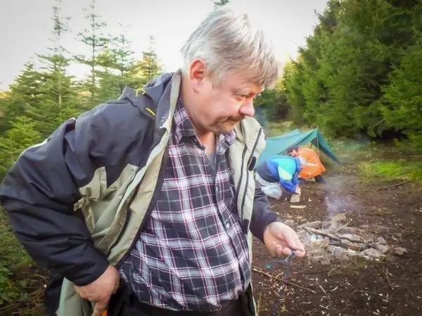 Mann hält einen Zunder in der Hand, während Rauch von einem kleinen Bodenfeuer aufsteigt