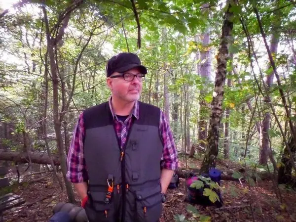 Mann in Wald mit Outdoor-Bekleidung und Ausrüstung, umgeben von Bäumen