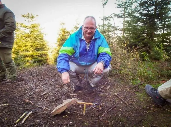 Mann kniet neben einer kleinen Flamme auf dem Boden, Holzstücke liegen verstreut