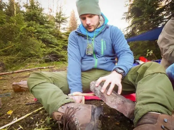 Mann sitzt im Freien und bearbeitet einen Holzstock mit einem Messer