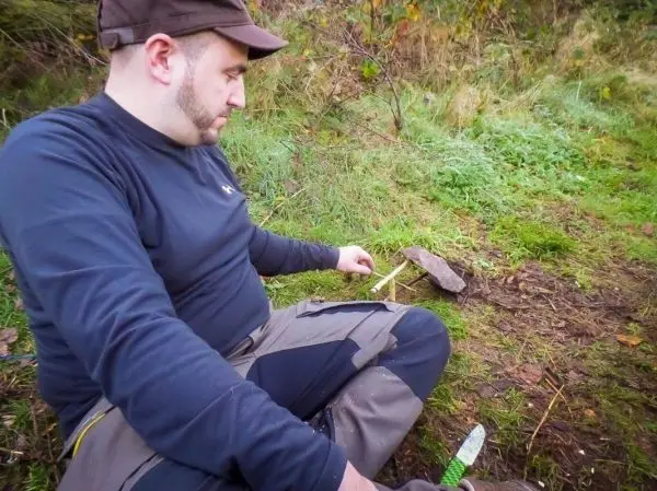 Mann sitzt im Gras und bearbeitet einen Stock mit einem Messer