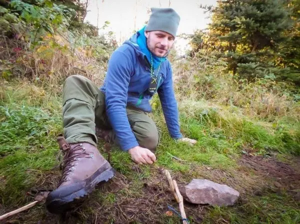 Mann sitzt im Gras und bearbeitet einen Stock mit einem Messer