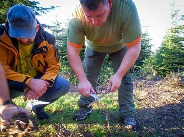 Messer schneidet Holzstück auf dem Boden, während zwei Personen beobachten