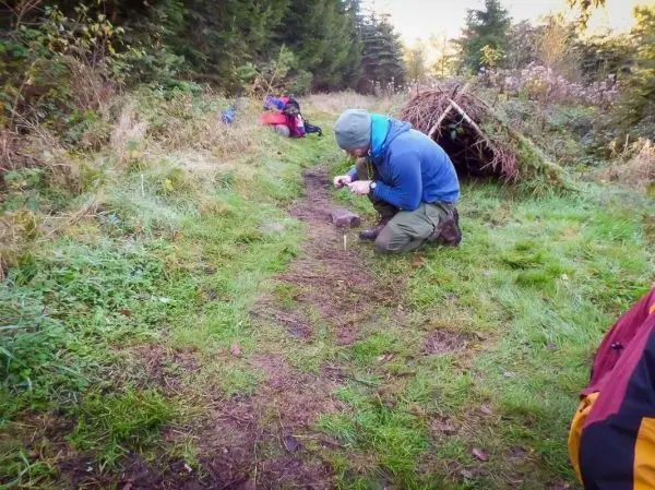 Messer schneidet Zunder auf einem Waldweg neben einer improvisierten Tarp-Notunterkunft