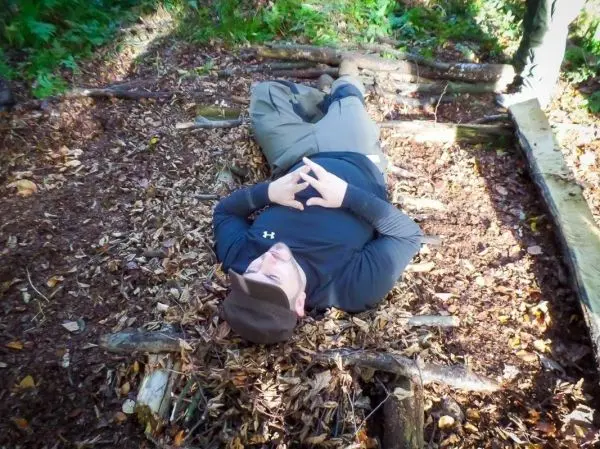 Person liegt auf einer improvisierten Liege aus Ästen und Laub im Wald