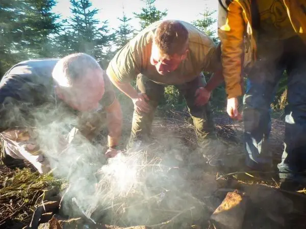 Zunder glimmt in einem kleinen Bodenfeuer, drei Personen beugen sich darüber