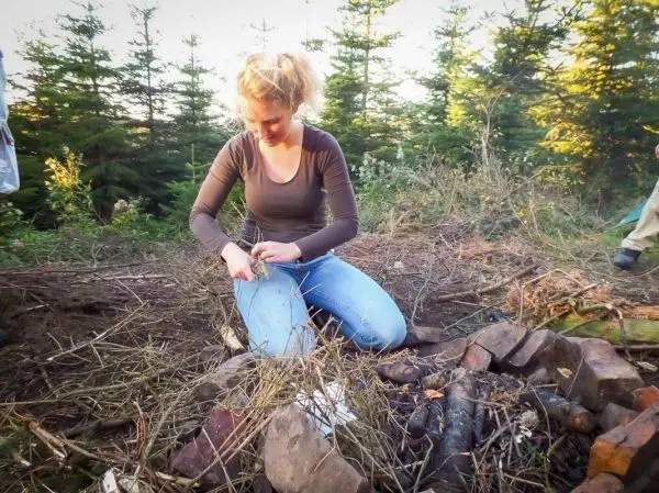 Zunder wird mit einem Messer bearbeitet, während ein Feuerplatz im Hintergrund sichtbar ist