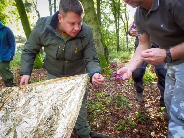 Zwei Personen spannen eine Tarp-Notunterkunft mit Schnüren zwischen Bäumen
