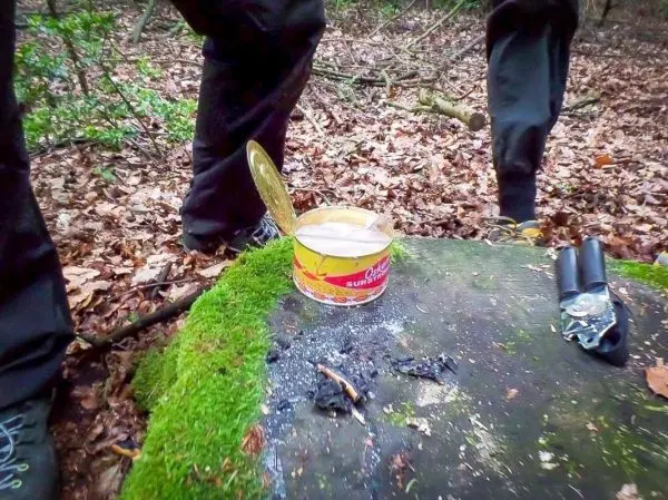 Dose mit geöffnetem Deckel steht auf einem moosbedeckten Stein, daneben ein Feuerstelle