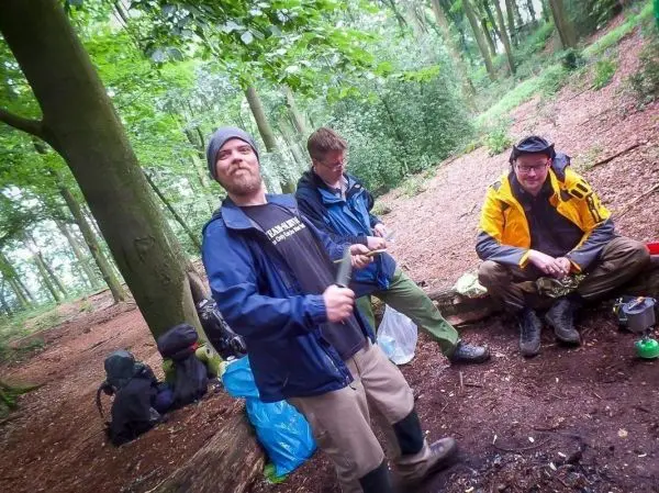 Drei Personen in einem Wald, eine hält ein Messer, andere sitzen am Boden