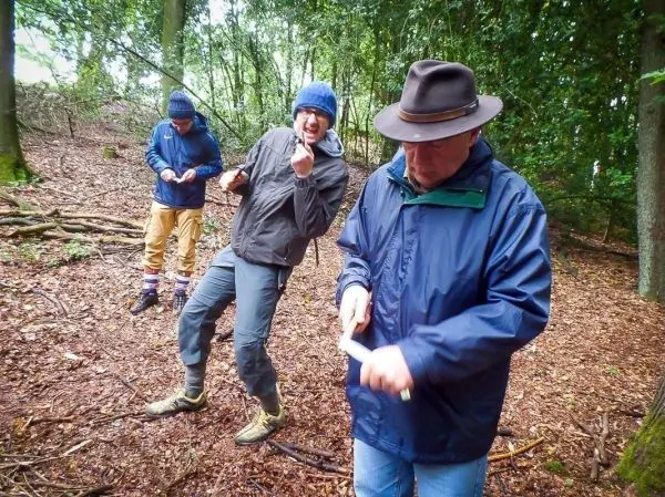 Drei Personen stehen im Wald und bearbeiten Holz mit Messern