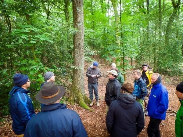 Gruppe diskutiert unter Bäumen im Wald, einige tragen Hüte und Jacken