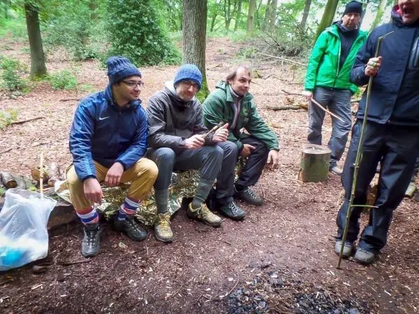 Gruppe sitzt auf einem Holzstamm neben einem kleinen Bodenfeuer im Wald