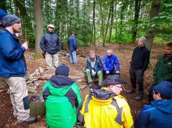 Gruppe sitzt auf Holzstämmen im Wald, einige Personen stehen um sie herum