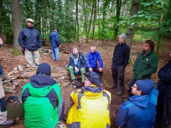 Gruppe sitzt im Wald um einen Holzstamm, diskutiert über Überlebenstechniken