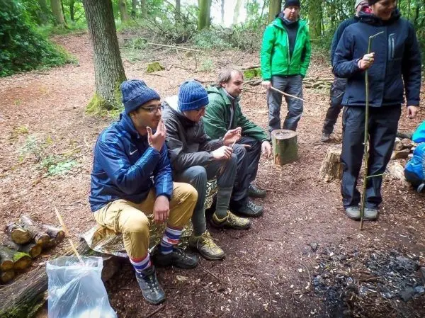 Gruppe sitzt um ein kleines Lagerfeuer, Holzstämme und Ausrüstung sichtbar