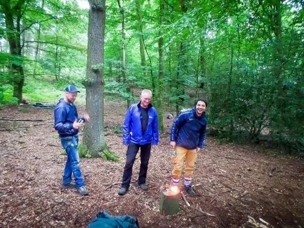 Kleines Bodenfeuer auf einem Baumstumpf im Wald, drei Personen stehen daneben
