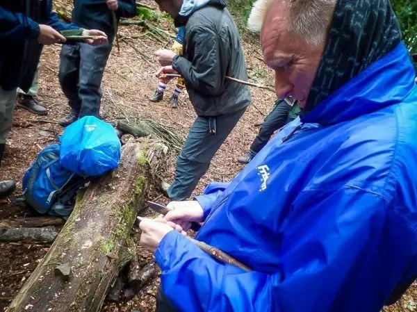 Mann bearbeitet Holzstück mit Messer, während andere im Hintergrund arbeiten