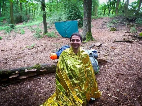 Mann in einer Rettungsdecke sitzt vor einem Rucksack und einer Tarp-Notunterkunft