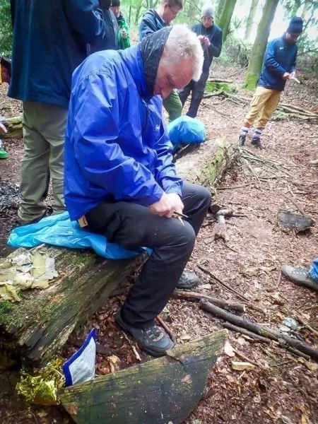 Mann sitzt auf einem Baumstamm und schnitzt mit einem Messer