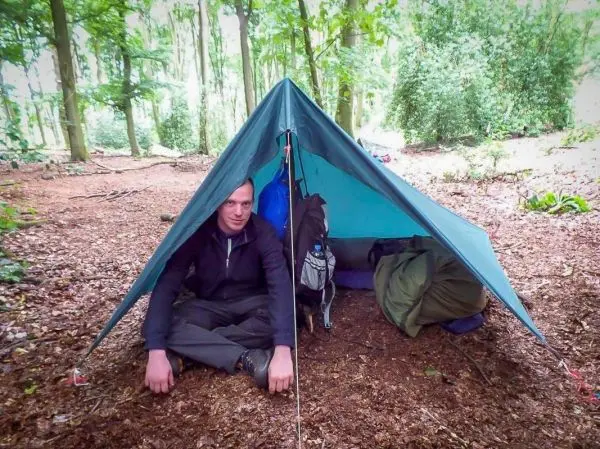 Mann sitzt vor einer Tarp-Notunterkunft im Wald, Rucksäcke liegen daneben
