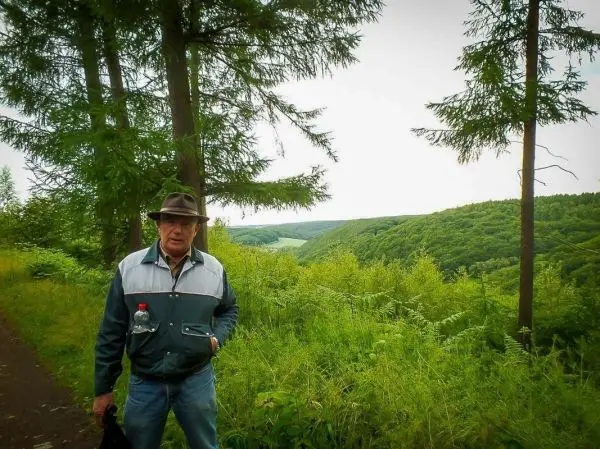 Mann steht auf einem Waldweg zwischen Bäumen mit Blick auf eine grüne Hügellandschaft