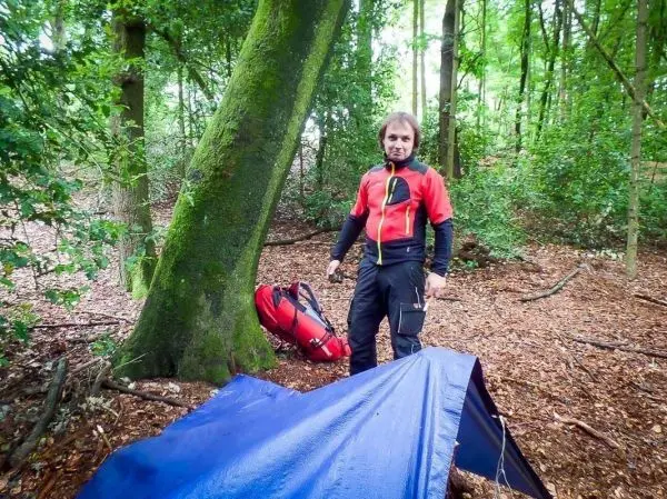 Mann steht neben einer blauen Tarp-Notunterkunft im Wald