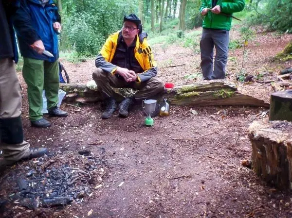 Person sitzt auf einem Baumstamm und beobachtet andere Personen beim Kochen im Wald
