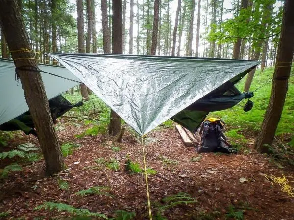 Tarp-Notunterkunft hängt zwischen Bäumen, darunter Rucksack und Holzunterlage