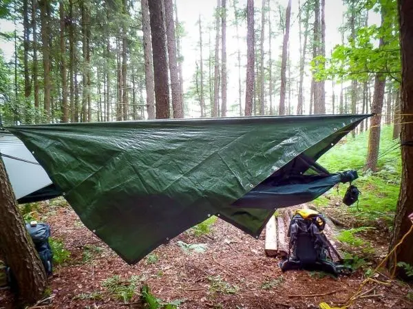 Tarp-Notunterkunft ist zwischen Bäumen gespannt, Rucksack liegt im Vordergrund
