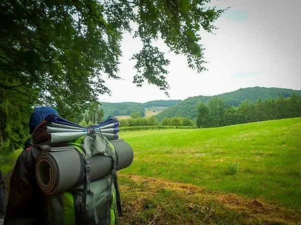 Wanderer mit Rucksack und Isomatten blickt auf eine grüne Wiese und Hügel