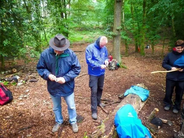 Zwei Personen bearbeiten Holzstücke im Wald, eine Person hält einen Stock
