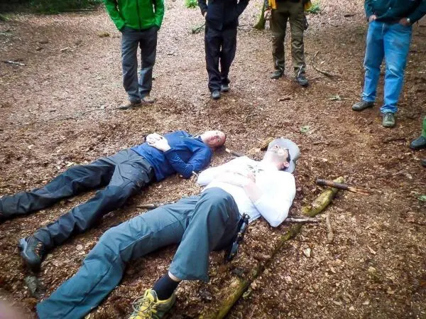 Zwei Personen liegen auf dem Waldboden, umgeben von mehreren stehenden Personen