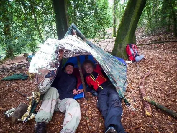 Zwei Personen sitzen in einer Tarp-Notunterkunft aus reflektierendem Material im Wald