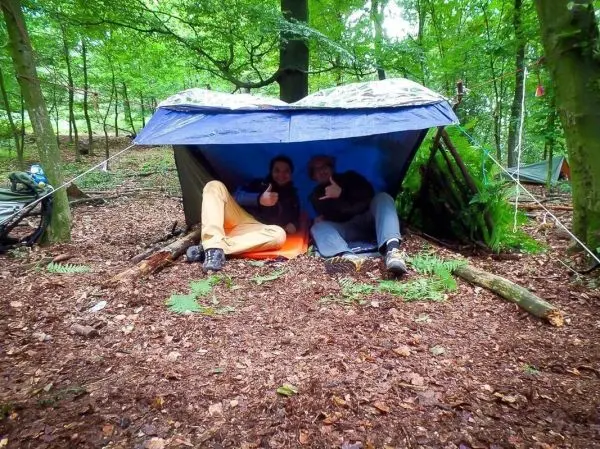 Zwei Personen sitzen unter einem selbstgebauten Unterschlupf im Wald