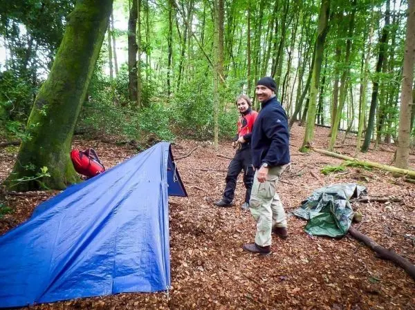 Zwei Personen stehen neben einer blauen Tarp-Notunterkunft im Wald