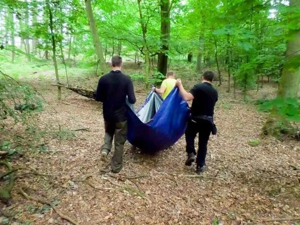 Drei Personen tragen eine improvisierte Trage durch den Wald