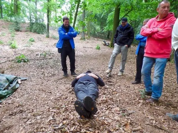 Gruppe beobachtet liegende Person auf dem Waldboden zwischen Bäumen und Laub
