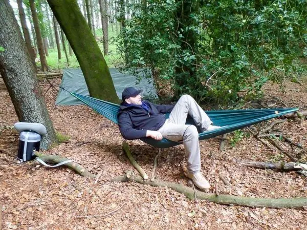 Hängematte zwischen Bäumen mit sitzender Person im Wald, Tarp im Hintergrund