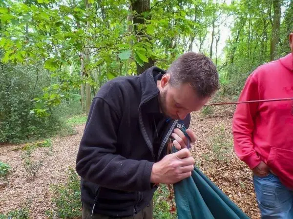 Mann befestigt eine Tarp-Notunterkunft mit einem Gestänge im Wald