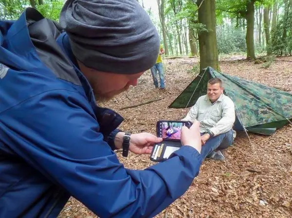 Mann fotografiert sitzenden Teilnehmer vor einer Tarp-Notunterkunft im Wald