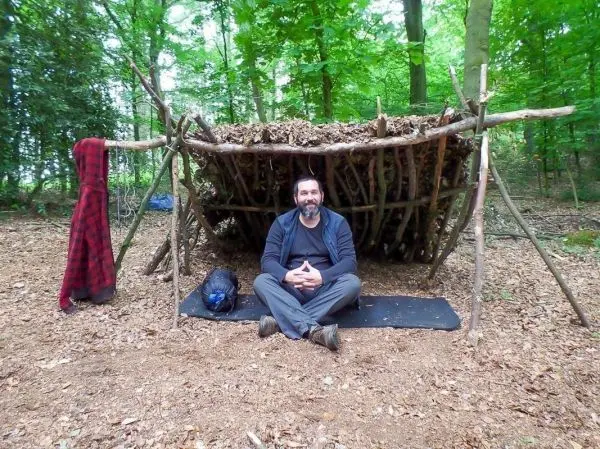 Mann sitzt vor einer selbstgebauten Laubhütte im Wald