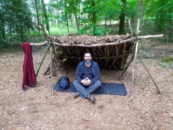 Mann sitzt vor einer selbstgebauten Laubhütte im Wald auf einer Matte