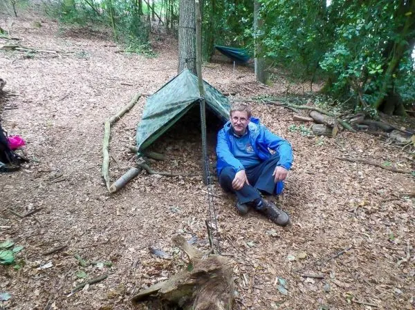Mann sitzt vor einer Tarp-Notunterkunft aus Plane und Ästen im Wald
