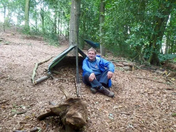 Mann sitzt vor einer Tarp-Notunterkunft im Wald auf dem Boden