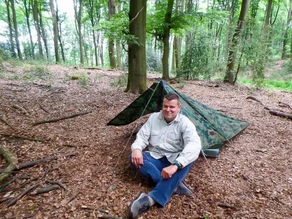 Mann sitzt vor einer Tarp-Notunterkunft im Wald auf Laub