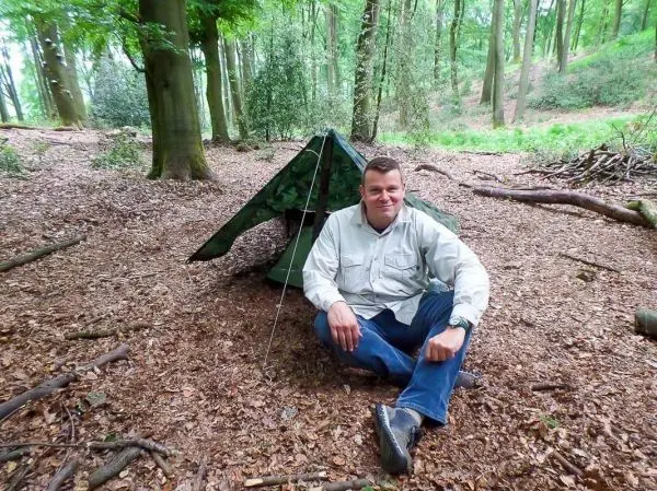 Mann sitzt vor einer Tarp-Notunterkunft im Wald auf Laub