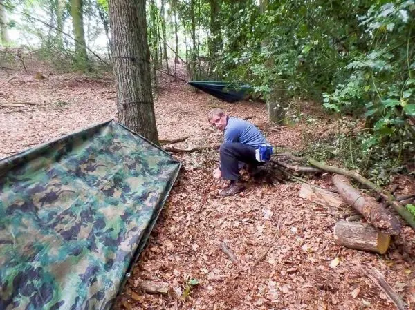 Person sitzt auf dem Boden neben einer camouflagefarbenen Plane im Wald