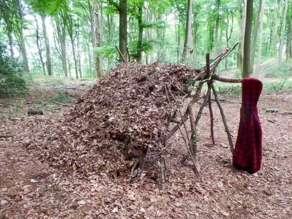Person steht neben einer selbstgebauten Laubhütte im Wald