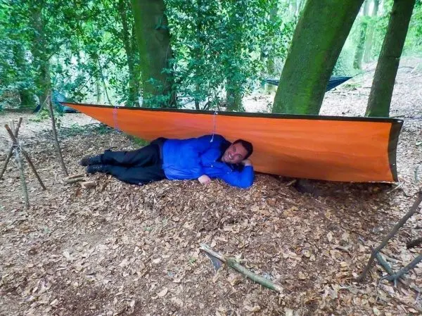 Tarp-Notunterkunft hängt zwischen zwei Bäumen, Person liegt darunter auf dem Boden